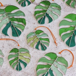 Peut inclure: Une collection d'ornements en verre color&eacute; en forme de feuilles de monstera avec des cordons orange. Les feuilles sont de diff&eacute;rentes nuances de vert.