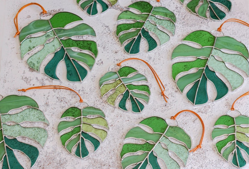 Peut inclure: Une collection d'ornements en verre color&eacute; en forme de feuilles de monstera avec des cordons orange. Les feuilles sont de diff&eacute;rentes nuances de vert.
