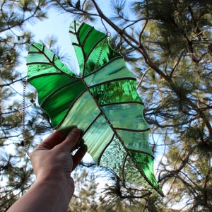 May include: A stained glass leaf with a green and white design, hanging from a silver chain. The leaf is held in front of a pine tree.