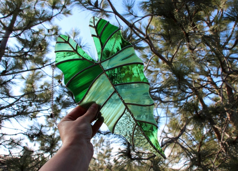 May include: A stained glass leaf with a green and white design, hanging from a silver chain. The leaf is held in front of a pine tree.