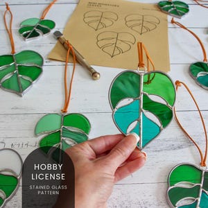 Peut inclure: Une collection d'ornements en verre teinté en forme de feuilles de monstera. Les feuilles sont faites de verre vert, bleu et blanc. Les ornements sont suspendus à des cordons bruns. Le texte "Hobby License Stained Glass Pattern" est imprimé sur un fond blanc.