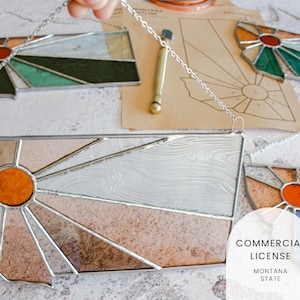 May include: Three stained glass sunburst designs in the shape of the state of Montana. The sunbursts are made with clear, white, orange, brown, blue, and green glass. The designs are hanging from silver chains.