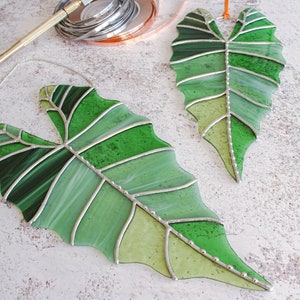 May include: Two stained glass leaves in shades of green, with silver accents and a chain for hanging. The leaves are shaped like a heart and have a detailed vein pattern.