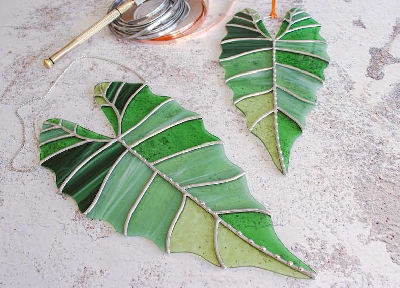 May include: Two stained glass leaves in shades of green, with silver accents and a chain for hanging. The leaves are shaped like a heart and have a detailed vein pattern.