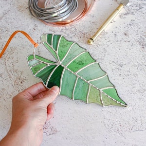 May include: A stained glass leaf in shades of green, with a brown leather cord attached. The leaf is a realistic depiction of a tropical plant.