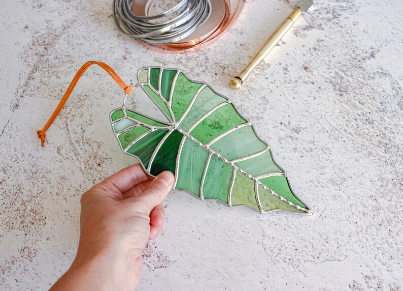 May include: A stained glass leaf in shades of green, with a brown leather cord attached. The leaf is a realistic depiction of a tropical plant.