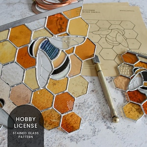 May include: A stained glass pattern for a beehive with a bee in the centre. The pattern is made of clear, yellow, and orange glass. The pattern is on a white background with a glass cutter and a piece of copper foil.