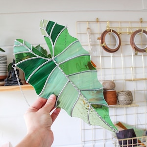 May include: A stained glass leaf in shades of green, with a silver chain attached to the top. The leaf is hanging from a gold metal grid.
