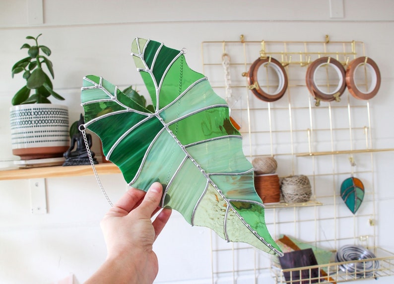 May include: A stained glass leaf in shades of green, with a silver chain attached to the top. The leaf is hanging from a gold metal grid.