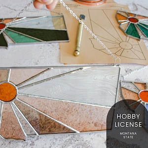 May include: A stained glass sunburst design in the shape of the state of Montana. The sunburst is made of various shades of brown, orange, and white glass. The glass is framed in silver metal and has a chain for hanging. The image also includes the text "Hobby License Montana State".