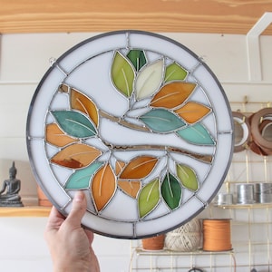 May include: A round stained glass window featuring a stylized branch with leaves in shades of green, orange, and brown. The leaves are arranged in a circular pattern around the branch.