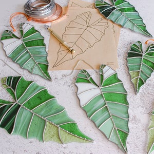 May include: Five stained glass leaf-shaped ornaments in shades of green and white. The leaves are arranged on a white surface.
