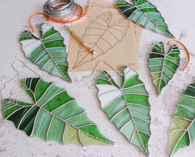 May include: Five stained glass leaf-shaped ornaments in shades of green and white. The leaves are arranged on a white surface.