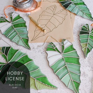 May include: Five stained glass leaf-shaped ornaments in shades of green and white, with silver foil accents. The ornaments are hanging from a white surface. The text "HOBBY LICENSE ALACACIA LEAF" is printed on a black rectangle in the lower center of the image.