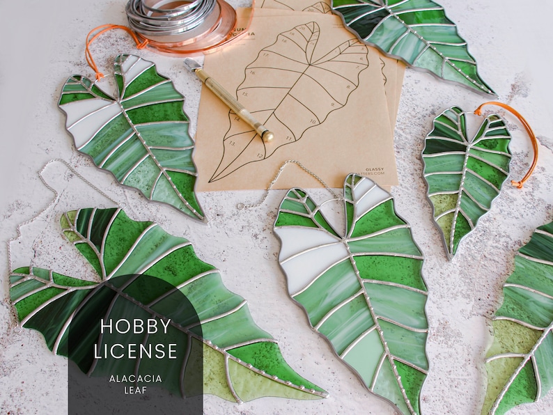 May include: Five stained glass leaf-shaped ornaments in shades of green and white, with silver foil accents. The ornaments are hanging from a white surface. The text "HOBBY LICENSE ALACACIA LEAF" is printed on a black rectangle in the lower center of the image.