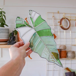 May include: A stained glass leaf in shades of green and white, with a brown leather cord attached. The leaf is hanging from a hand.