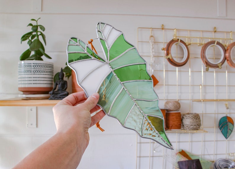 May include: A stained glass leaf in shades of green and white, with a brown leather cord attached. The leaf is hanging from a hand.