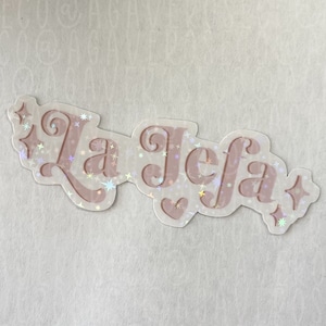 May include: A white sticker with pink glitter and the text "La Jefa" in a pink script font.