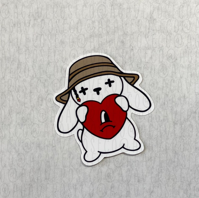 Może przedstawiać: Biała naklejka przedstawiająca kresk&oacute;wkowego kr&oacute;lika w brązowym kapeluszu, trzymającego czerwone serce ze smutną miną. Kr&oacute;lik ma czarne oczy w kształcie krzyża i papierosa w ustach.