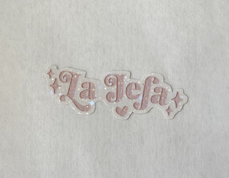 May include: A clear sticker with the text "La Jefa" in pink script with a glittery effect. The sticker has a white background and is surrounded by small stars.