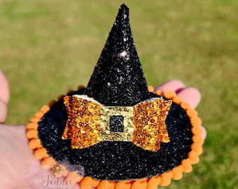 Puede incluir: Un pequeño sombrero de bruja negro con purpurina, con un lazo naranja con purpurina y una hebilla dorada. El sombrero tiene un ribete de pompón naranja en la base. Accesorio temático de Halloween.