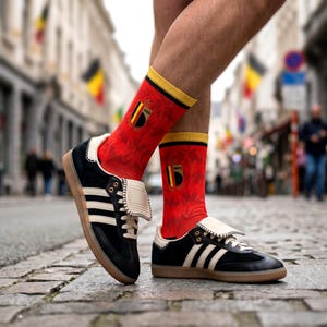 May include: Black and white trainers with gum soles and folded tongues paired with red crew socks featuring a yellow and black vertical stripe emblem and the text 1895. The socks have a yellow top band and a subtle tonal pattern throughout the fabric.
