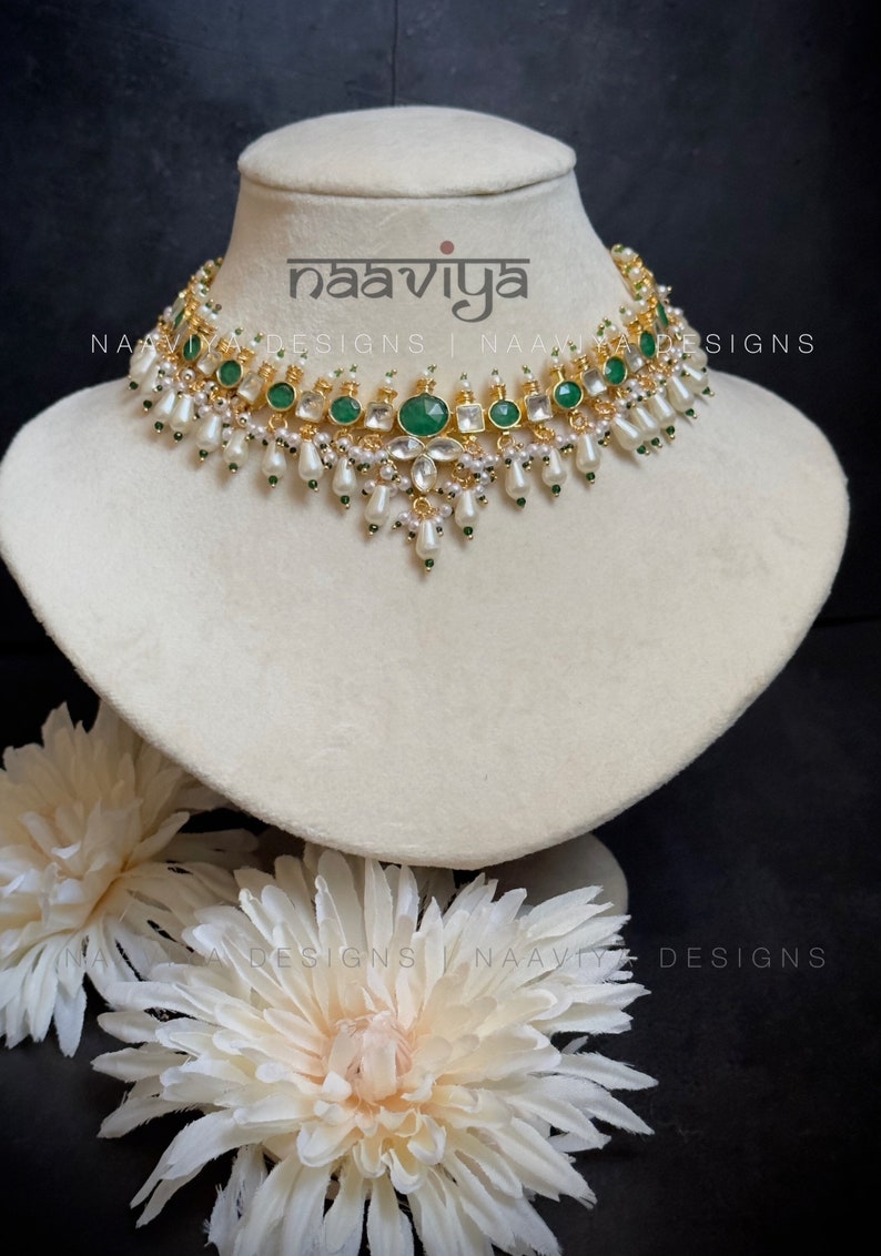 K&ouml;nnte beinhalten: Eine goldene und gr&uuml;ne Choker-Kette mit wei&szlig;en Perlen und smaragdgr&uuml;nen Steinen. Die Kette befindet sich auf einer wei&szlig;en Mannequin-B&uuml;ste. Der Text "naaviya designs" ist auf der B&uuml;ste sichtbar.