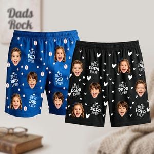 May include: Two pairs of athletic shorts, one blue and one black, featuring a repeating pattern of children's faces, small crowns, and text. The blue shorts display baseballs and the text BEST DAD EVER, while the black shorts feature white hearts and the text BEST PAPA EVER.