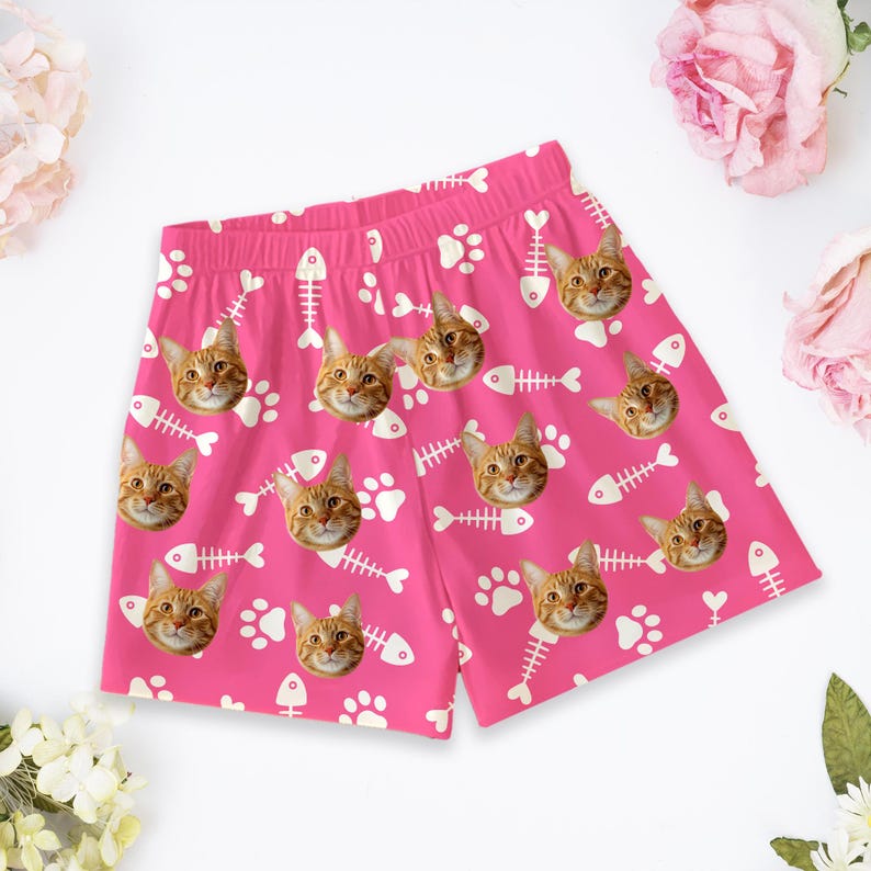 K&ouml;nnte beinhalten: Hellrosa Shorts mit einem durchgehenden Muster aus orangefarbenen getigerten Katzengesichtern, wei&szlig;en Fischgr&auml;tensilhouetten und wei&szlig;en Pfotenabdr&uuml;cken. Die Shorts verf&uuml;gen &uuml;ber einen elastischen Bund und eine lockere Passform.