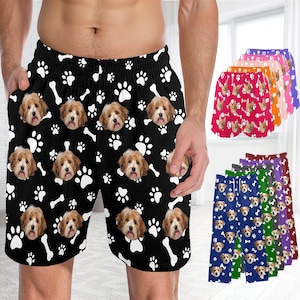 K&ouml;nnte beinhalten: Schwarze Shorts mit einem sich wiederholenden Muster aus wei&szlig;en Hundepfotenabdr&uuml;cken, wei&szlig;en Knochen und dem Gesicht eines hellbraunen Hundes. Die Shorts haben einen elastischen Bund und Seitentaschen. Weitere Farbvarianten sind im Hintergrund zu sehen.