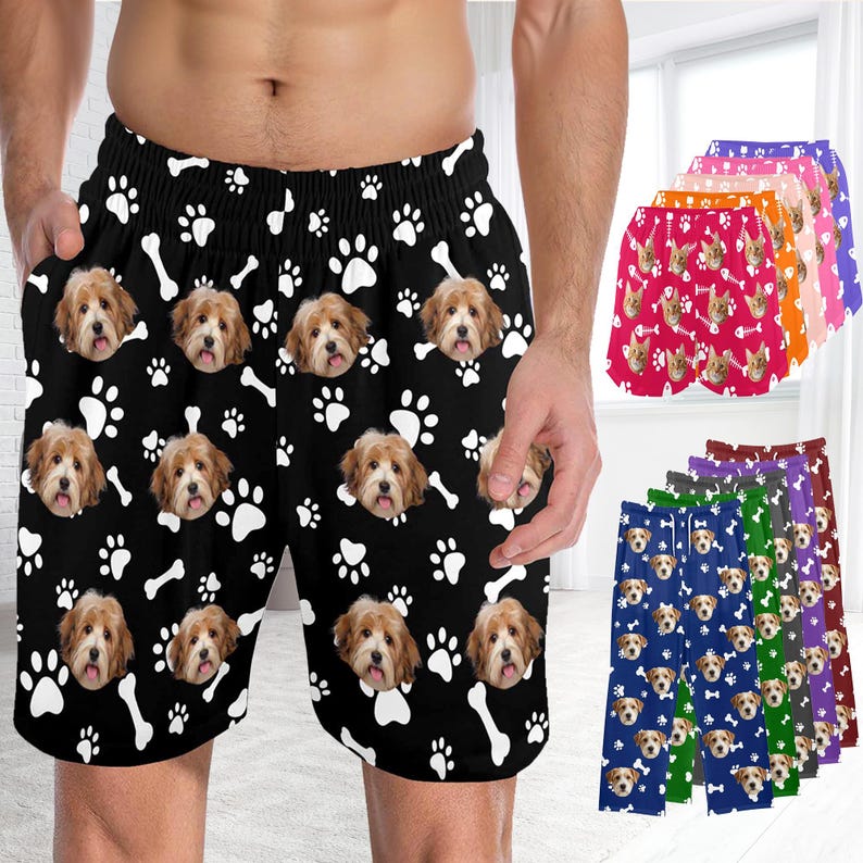 K&ouml;nnte beinhalten: Schwarze Shorts mit einem sich wiederholenden Muster aus wei&szlig;en Hundepfotenabdr&uuml;cken, wei&szlig;en Knochen und dem Gesicht eines hellbraunen Hundes. Die Shorts haben einen elastischen Bund und Seitentaschen. Weitere Farbvarianten sind im Hintergrund zu sehen.