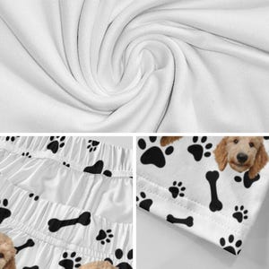 K&ouml;nnte beinhalten: Wei&szlig;er Stoff mit einem sich wiederholenden Muster aus schwarzen Hundepfotenabdr&uuml;cken und schwarzen Knochen. Das Design enth&auml;lt eine Teilansicht eines hellbraunen Hundegesichts mit Schlappohren. Das Material zeigt Falten und sichtbare N&auml;hte.