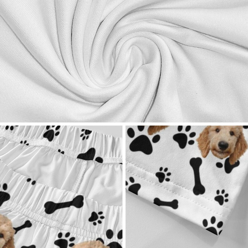 K&ouml;nnte beinhalten: Wei&szlig;er Stoff mit einem sich wiederholenden Muster aus schwarzen Hundepfotenabdr&uuml;cken und schwarzen Knochen. Das Design enth&auml;lt eine Teilansicht eines hellbraunen Hundegesichts mit Schlappohren. Das Material zeigt Falten und sichtbare N&auml;hte.