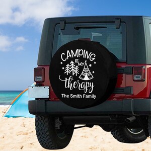 May include: Black spare tire cover with white text that reads "Camping is my therapy" with a stylized image of a tent and trees. The text "The Smith Family" is below the main text.