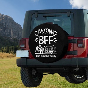 May include: A black spare tire cover with white text that reads "Camping BFF The Smith Family". The cover features a small camper van and pine trees. The cover is mounted on a red Jeep.
