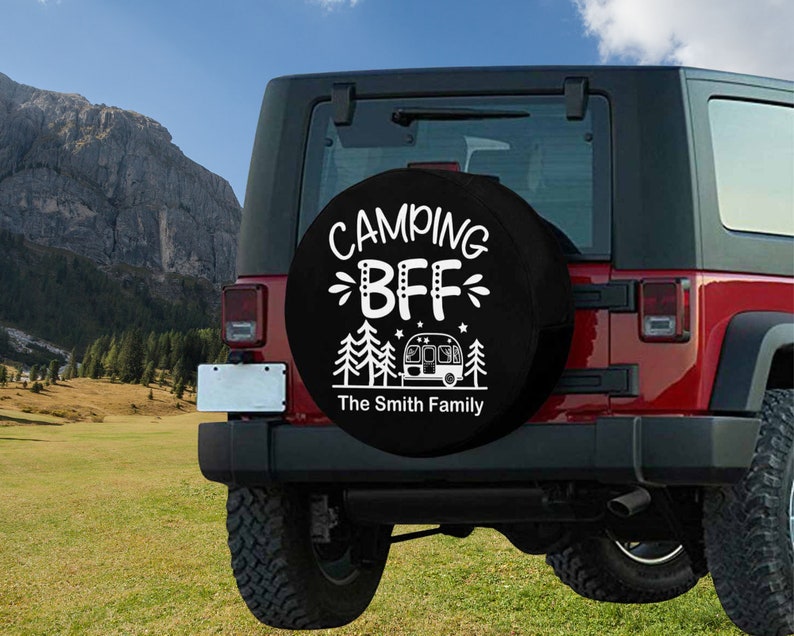 May include: A black spare tire cover with white text that reads "Camping BFF The Smith Family". The cover features a small camper van and pine trees. The cover is mounted on a red Jeep.