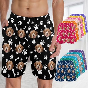 May include: Black shorts with a repeating pattern of white paw prints, dog faces, and bones. Additional shorts in various colors with similar dog-themed patterns are displayed. The shorts appear to be made of a lightweight fabric.