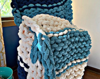 Puede incluir: Una manta de punto grueso en color crema, azul verdoso y azul marino. La manta está hecha de hilo suave y tiene una textura esponjosa.