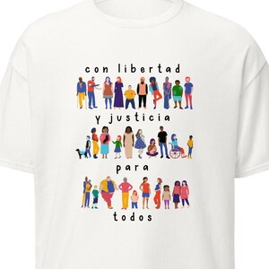 May include: White t-shirt with the Spanish text "con libertad y justicia para todos" in black. The shirt features colorful illustrations of diverse people. The design includes figures of various sizes, skin tones, and abilities.