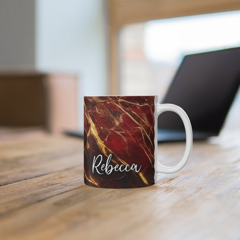 May include: White ceramic mug with a red and gold marble design. The mug has the name "Rebecca" written in white cursive on the front.