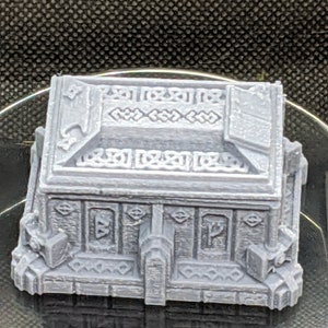 May include: A gray, intricately detailed, 3D printed treasure chest with a hinged lid and decorative carvings. The chest has a raised base with intricate details and runes.
