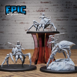 Op de afbeelding: Drie 3D-geprinte miniaturen van vrouwelijke figuren die spinnenachtige wezens berijden. De figuren zijn in lichtgrijs geverfd en staan in dynamische actiehoudings. De miniaturen staan op zwarte ronde bases.