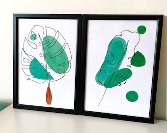 Könnte beinhalten: Zwei gerahmte Drucke mit schwarzen Rahmen. Die Drucke zeigen Linienzeichnungen von grünen Blättern mit Aquarell-Waschungen in Grüntönen. Der linke Druck zeigt ein Monstera-Blatt, während der rechte Druck ein einzelnes Blatt mit einem Stiel zeigt.