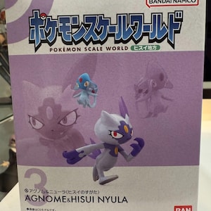 May include: A box featuring the "Pokémon Scale World" series, with the text "AGNOME & HISUI NYULA." The box has a purple and white color scheme, with illustrations of Pokémon characters. The Bandai Namco logo is visible.