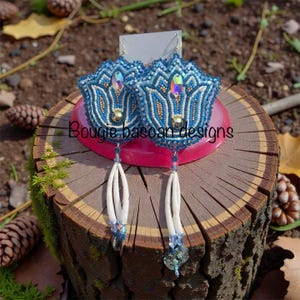 Può includere: Un paio di orecchini di perline blu con un design floreale. Gli orecchini hanno dettagli dorati e bianchi, con una gemma iridescente al centro. Da ogni orecchino pendono nappe bianche. Il testo "Bougie basoan designs" è visibile.