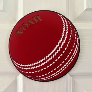 May include: Red cricket ball door hanger with white stitching and the name "NOAH" cut out in the centre.