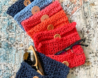 May include: A stack of six crocheted eyeglass cases in various shades of blue, orange, and red. Each case has a wooden button closure. The top case has a pair of sunglasses inside.