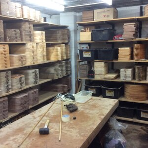 Op de afbeelding: Een werkplaats met planken vol houten snijplanken die in verschillende maten en vormen zijn gestapeld. De planken zijn gemaakt van verschillende soorten hout, waaronder esdoorn, kers en walnoot. Op de werkbank op de voorgrond liggen ook wat gereedschap en benodigdheden.