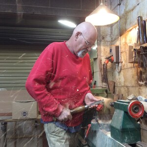 Op de afbeelding: Een man in een rood shirt gebruikt een houtdraaiinstrument om een ​​stuk hout op een draaibank te vormen. Houtspaanders vliegen van het draaiende hout.