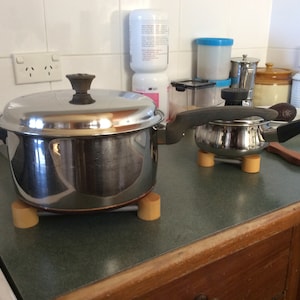 May include: Two stainless steel saucepans with lids and handles are on a kitchen counter. They are raised on small, light-coloured wooden trivets. Kitchen equipment.